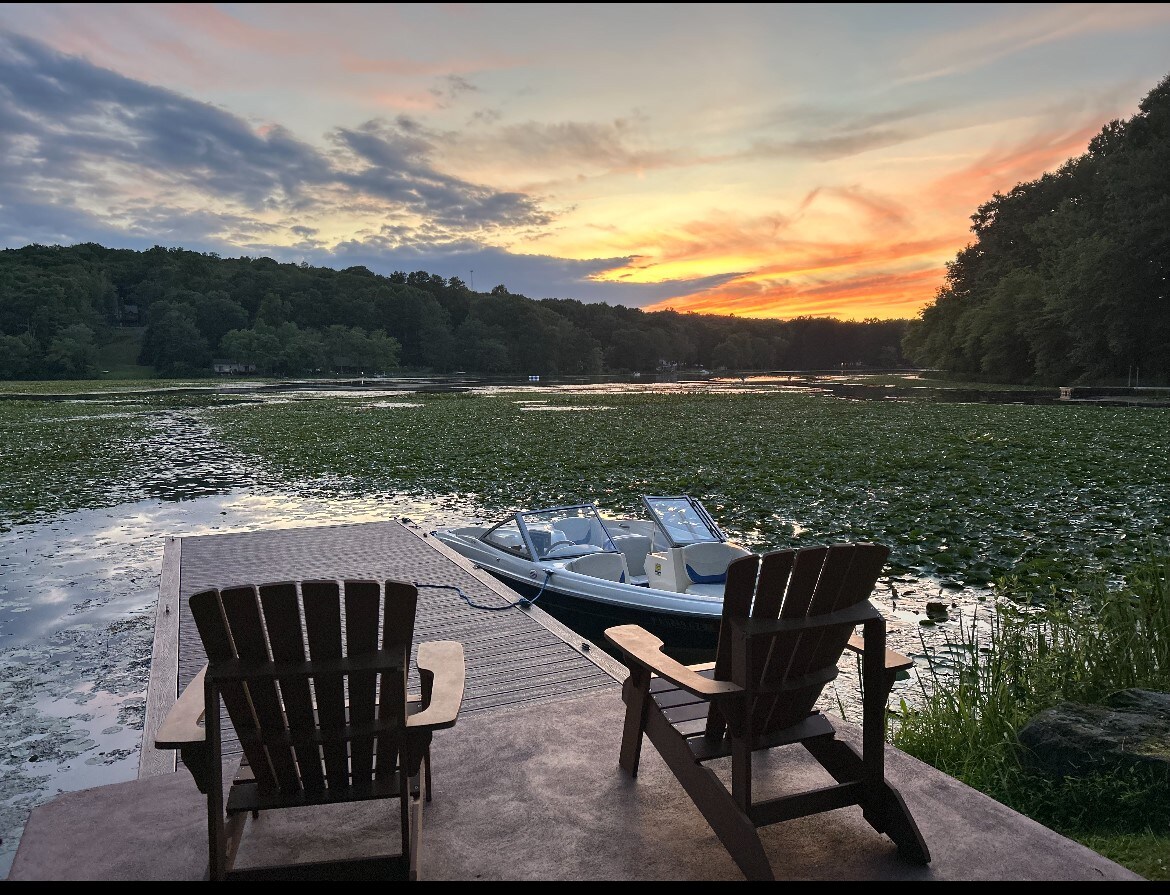 Two Adirondack chairs are positioned on a dock overlooking a calm lake. A small boat is tied to the dock, with lush green lily pads covering parts of the water. A vibrant sunset casts warm colors across the sky, providing a serene atmosphere.