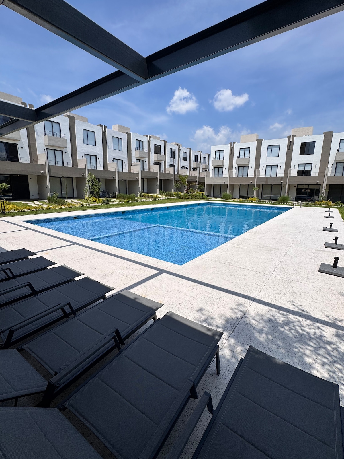 A rectangular swimming pool is centrally located, surrounded by a paved deck and lounge chairs. The area is framed by modern buildings featuring large windows. Clear blue skies and soft clouds provide a serene backdrop to this inviting outdoor space.