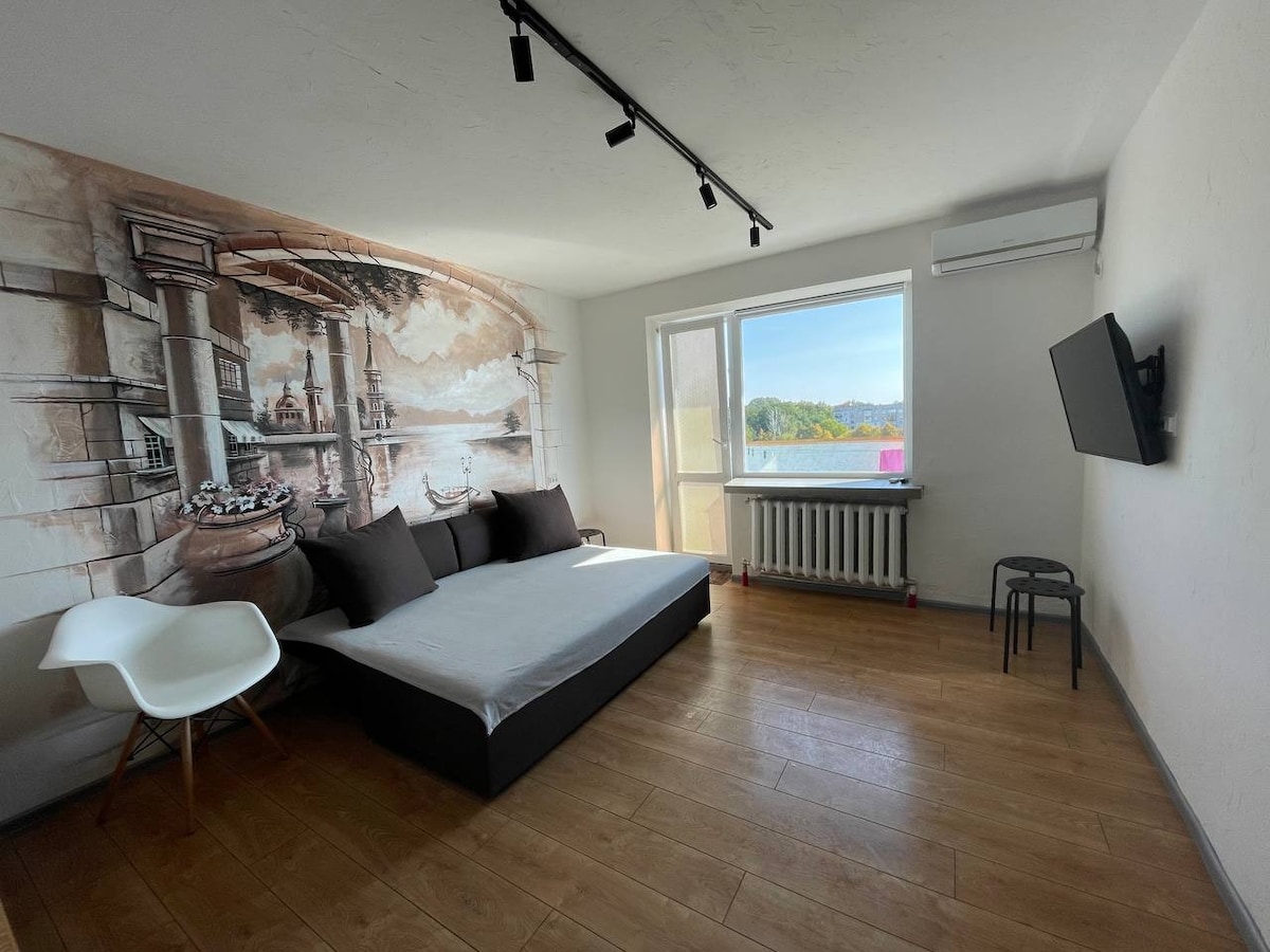 The living area features a minimalist design with a light-colored wall mural depicting a scenic landscape. A spacious, unmade sofa bed is positioned nearby, complemented by a modern chair and a small black side table. Large windows provide natural light and views of the outside.