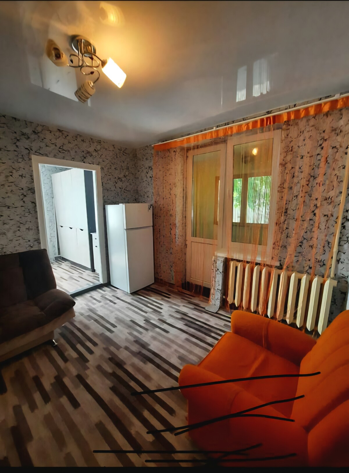 A modest living space features a bright orange sofa and a plush gray couch set on patterned flooring. Natural light filters through sheer curtains, illuminating the area. A white refrigerator stands against one wall, while a closet door is visible in the background.
