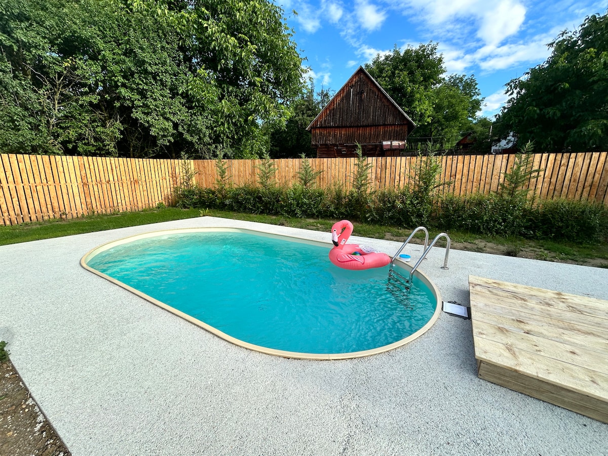 A private pool with a lightly curved shape is surrounded by a smooth deck. A pink inflatable float rests on the water's surface. Tall greenery and a wooden fence frame the pool area, creating a natural ambiance along with a glimpse of a wooden structure in the background.