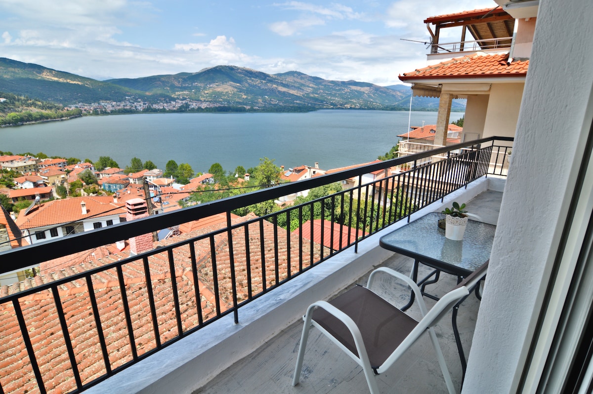 A balcony offers a panoramic view of the lake and surrounding hills, with terracotta rooftops visible below. Two outdoor chairs are arranged beside a small table, creating a comfortable space to enjoy the scenery. The sky is bright and partly cloudy.