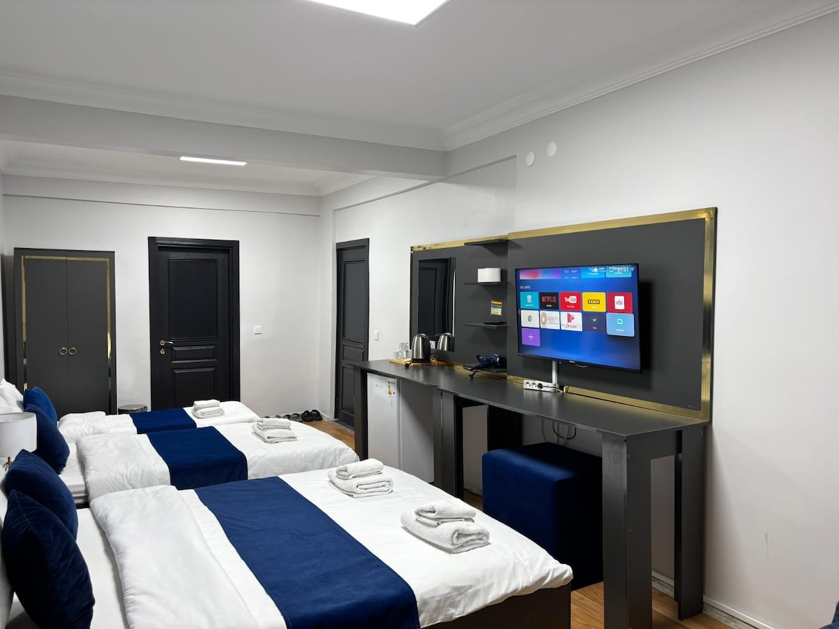 A hotel room contains two spacious beds dressed in white linens with blue accents. A sleek entertainment unit is mounted on the wall, featuring a large screen and modern amenities. Light reflects off the minimalist decor, enhancing the overall bright atmosphere.
