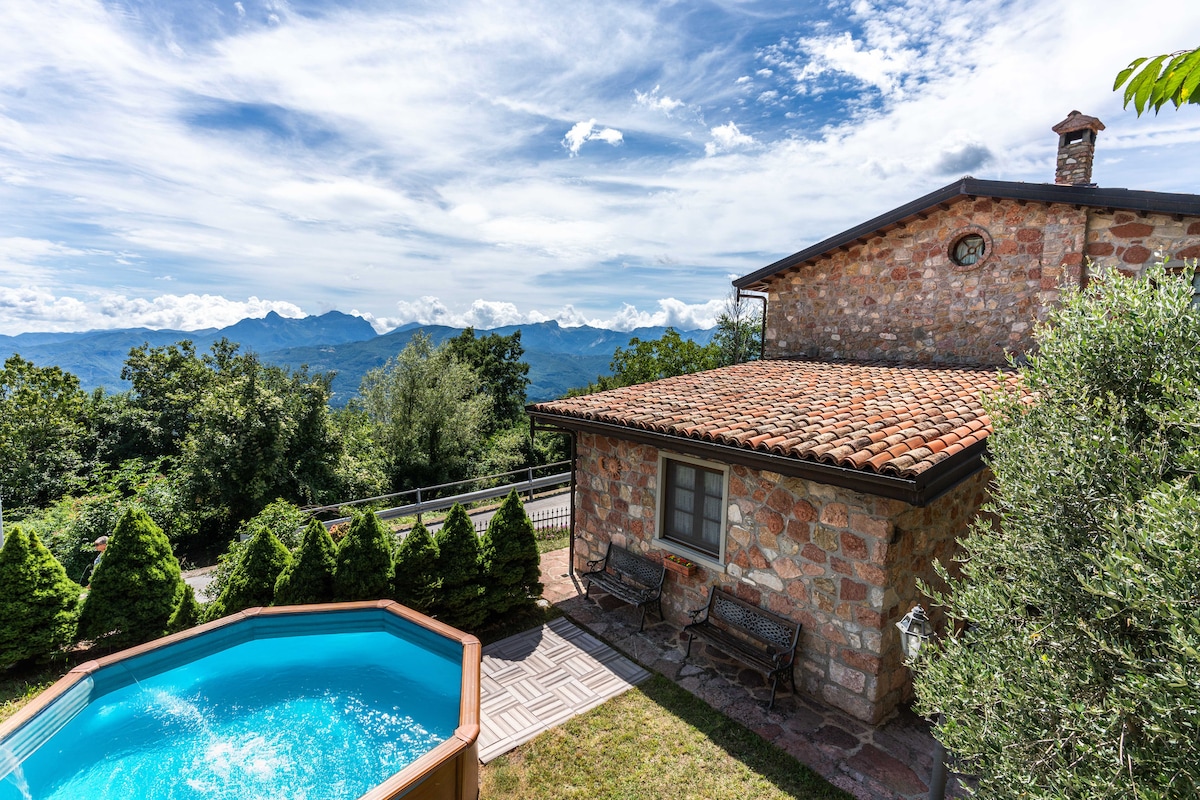 A stone house with a terracotta roof is surrounded by greenery, featuring a private above-ground pool in the foreground. The scenic mountain view stretches across the horizon under a partly cloudy sky, providing a peaceful setting for relaxation.