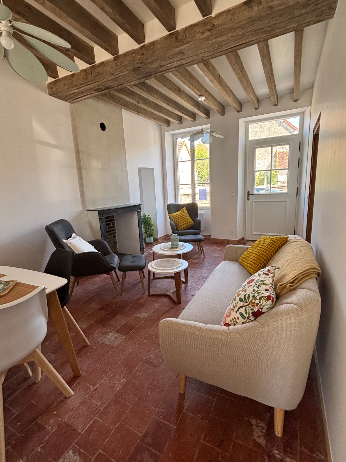 A living area features a cozy sofa with decorative cushions, complemented by two armchairs. A small round table is positioned in front, and a fireplace adds a touch of warmth. Large windows allow natural light, while exposed wooden beams enhance the character of the space.