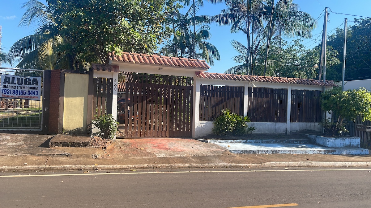 A view of a charming exterior featuring a wooden fence within a tropical setting. Lush greenery surrounds the property, with palm trees lining the area. A pathway leads to a gated entrance, providing a sense of privacy and tranquility.