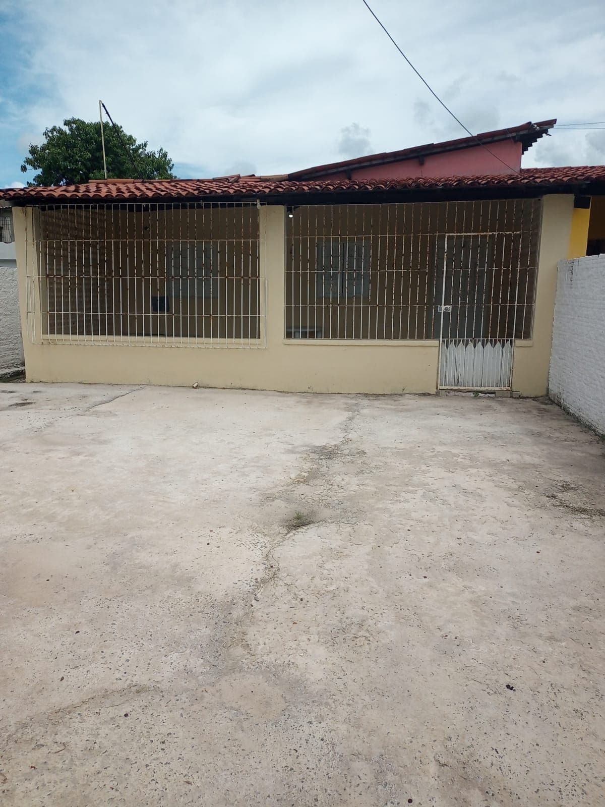 A single-story house is presented with a gated entrance, featuring a front courtyard with a concrete floor. Large, gridded windows allow natural light to filter into the interior, and the roof is covered with clay tiles, providing a distinct architectural element.