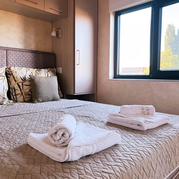 A spacious bed is dressed with a quilted cover and decorative pillows. Fresh towels are neatly arranged on the bed. Natural light enters the room through a large window, complementing the calming wall colors and simple furnishings.