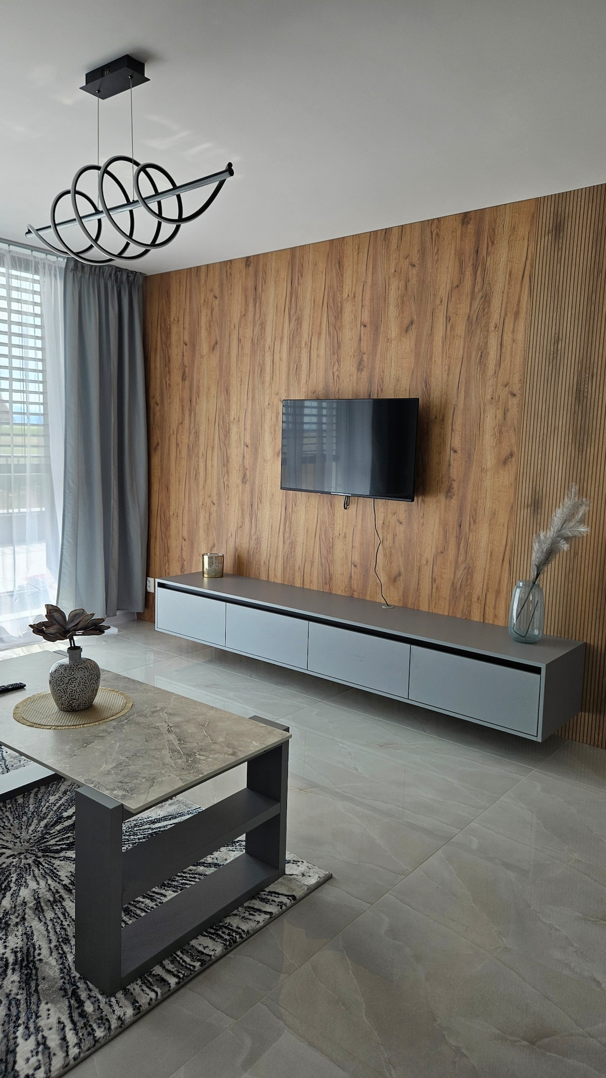 A modern living area features a sleek TV mounted on a wooden accent wall. A minimalist coffee table with a marble top sits on a textured rug. Light-filtering curtains soften the natural light streaming through the windows, creating a calming environment.