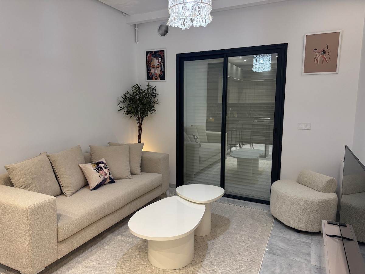 A modern living area is displayed, featuring a light-colored sofa adorned with a decorative pillow. Two white coffee tables sit in front of the sofa on a neutral rug. A sliding glass door brings in natural light, while a small decorative plant adds a touch of greenery.