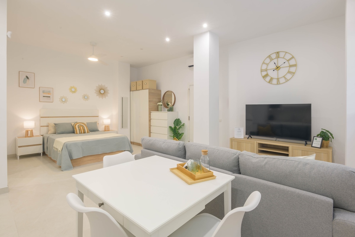 An open living space features a spacious bed with light bedding and decorative pillows, complemented by a stylish light fixture above. A comfortable sofa and dining table are visible in the foreground, while a modern TV unit and wall clock add functional detail to the cozy environment.