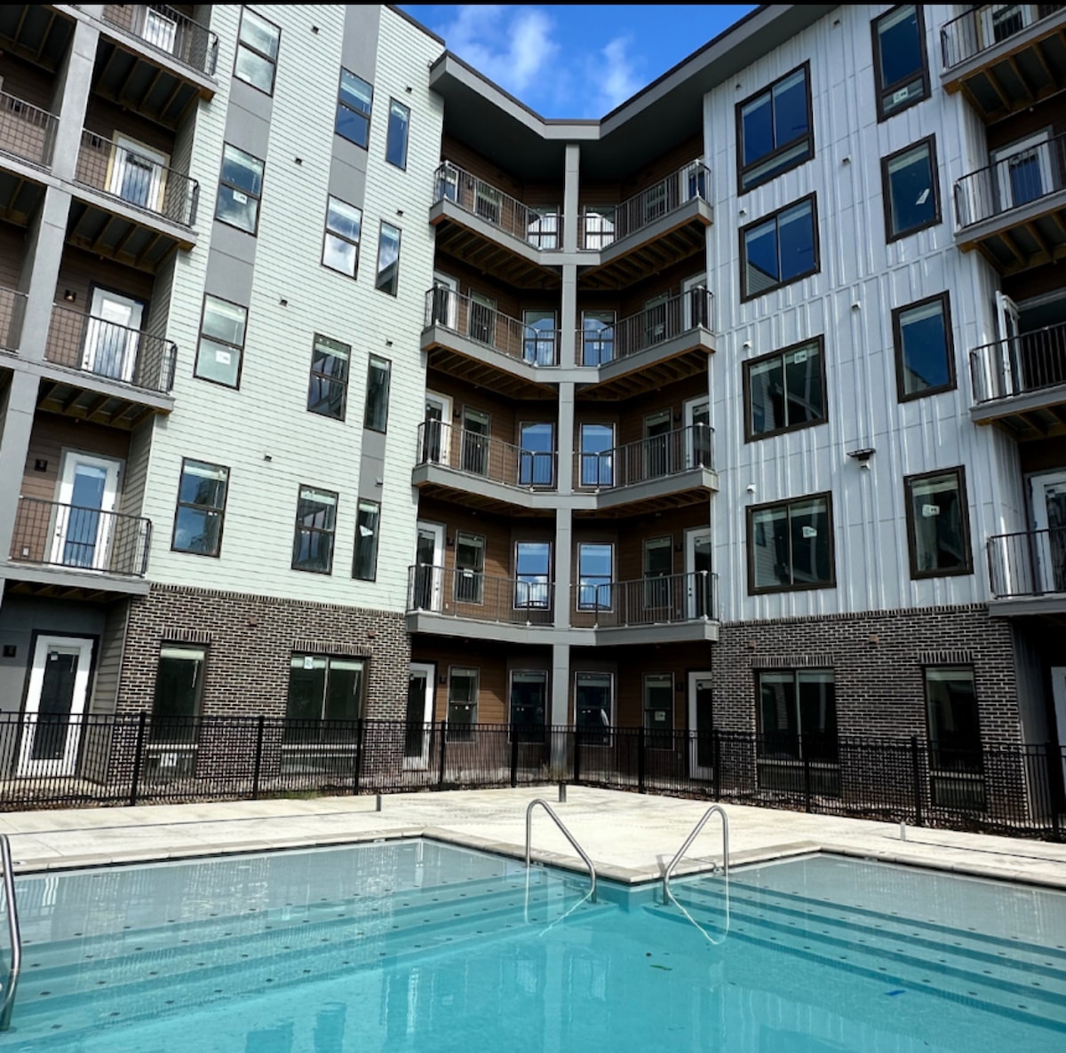 An outdoor swimming pool is located in an enclosed courtyard surrounded by a modern multi-story building. Several balconies are visible on the building's exterior, with large glass windows reflecting the sky. A pool ladder is positioned at one end of the pool.