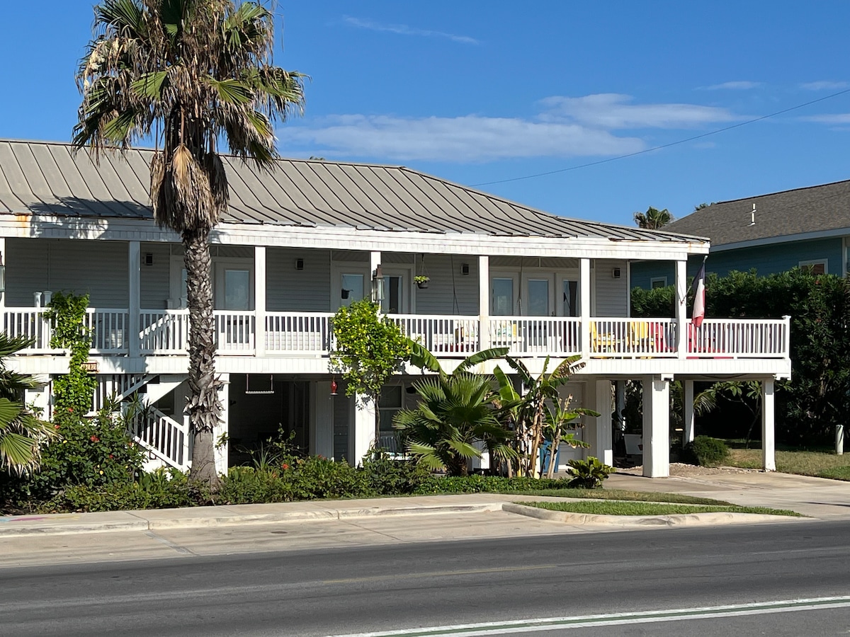 A light-colored building with a balcony is showcased against a clear blue sky. Palm trees and greenery are positioned in front, adding to the coastal appeal. The structure features multiple entrances and outdoor seating areas, creating a welcoming sight for guests.