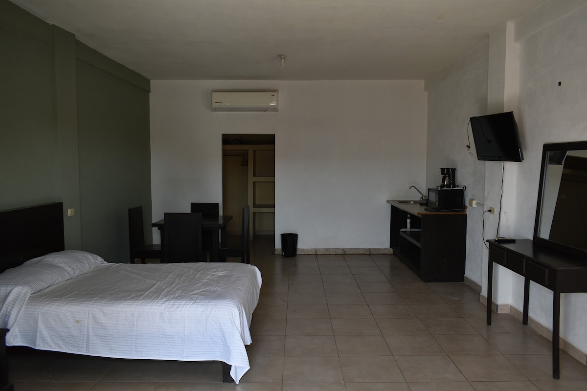 An open space is shown, featuring a spacious bed with a white coverlet. A small dining area with a table and chairs is located in one corner. A kitchenette is visible, equipped with essential appliances. A sizable mirror and a TV are mounted on the wall.
