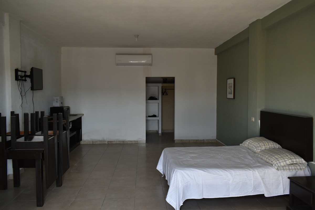 A spacious room has a bed with a white bedspread and a pillow on display. A simple dining table and chairs are positioned nearby. The kitchenette features essential appliances, and a closet is visible in the background. Natural light enters from a nearby window.