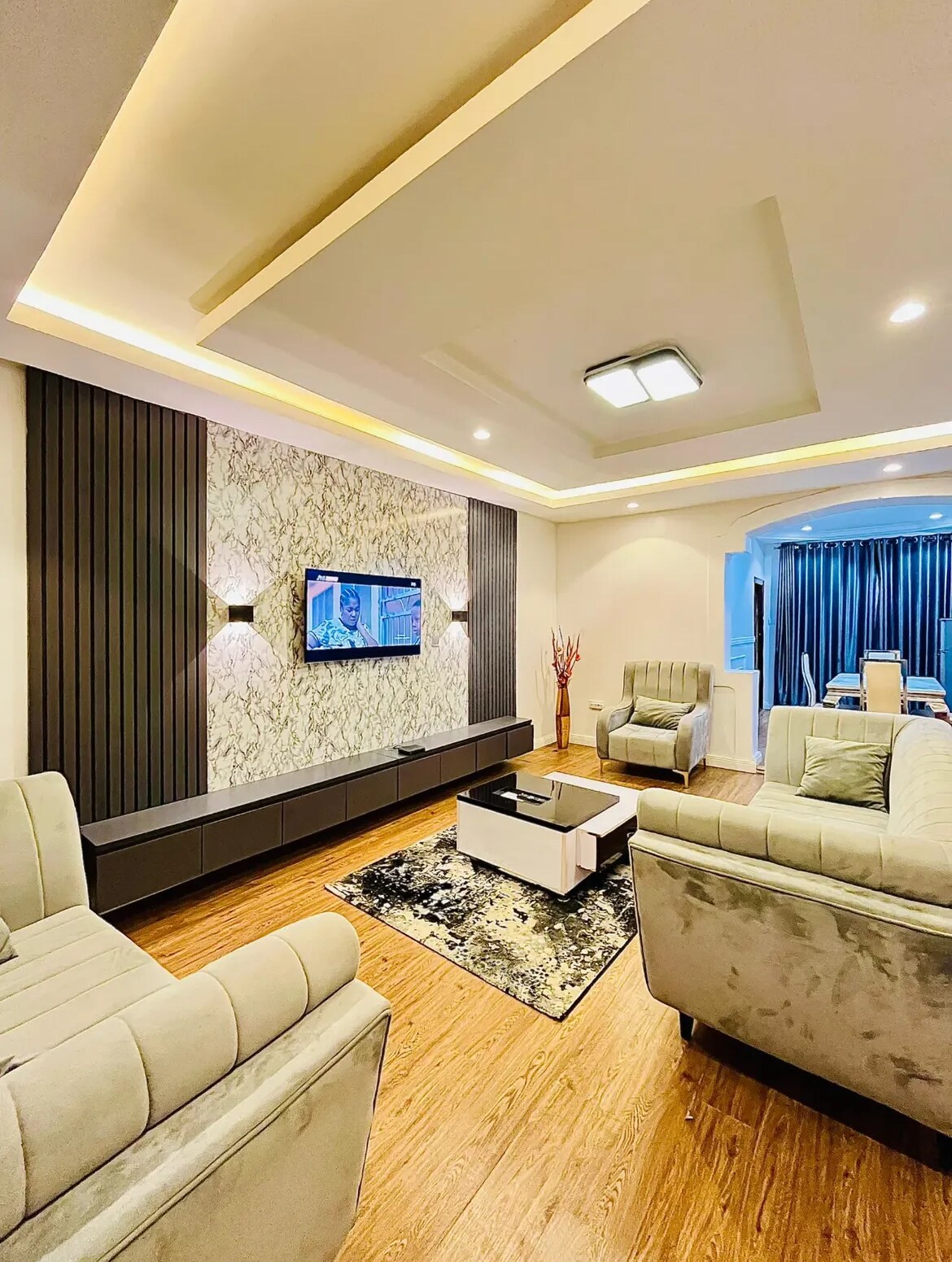 A spacious living area is highlighted by a stylish wall featuring decorative elements and mounted television. Comfortable seating is arranged around a central coffee table, while a patterned rug adds texture to the wooden floor. Soft lighting enhances the welcoming ambiance.