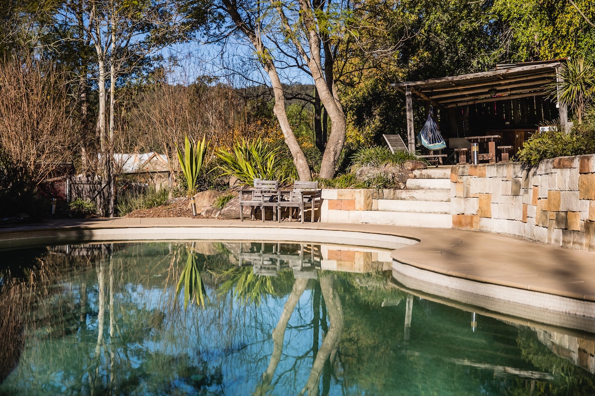 A serene swimming pool is framed by stone steps and lush greenery. A covered outdoor lounge area is visible nearby, featuring wooden furniture. The surrounding plants and trees reflect in the clear water, enhancing the peaceful atmosphere of the space.