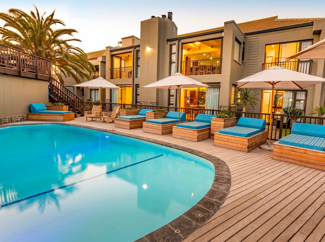 A refreshing swimming pool is surrounded by stylish lounge chairs with blue cushions, inviting relaxation. Umbrellas provide shade for outdoor seating areas. The two-story resort building features multiple balconies, enhancing the open and airy environment, with palm trees providing a tropical backdrop.