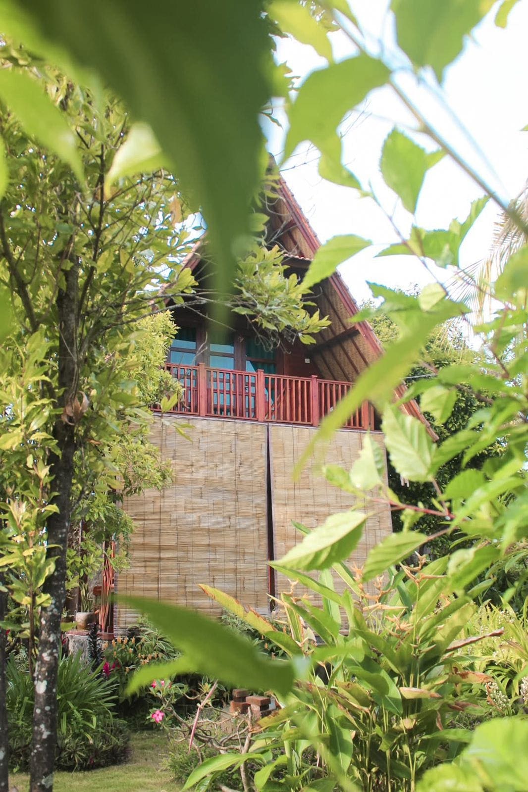 The traditional wooden cottage is partially obscured by lush green foliage, showcasing its elevated structure. A spacious balcony is visible, adorned with wooden railings. Surrounding tropical plants and trees enhance the natural setting, creating a serene atmosphere within the garden.