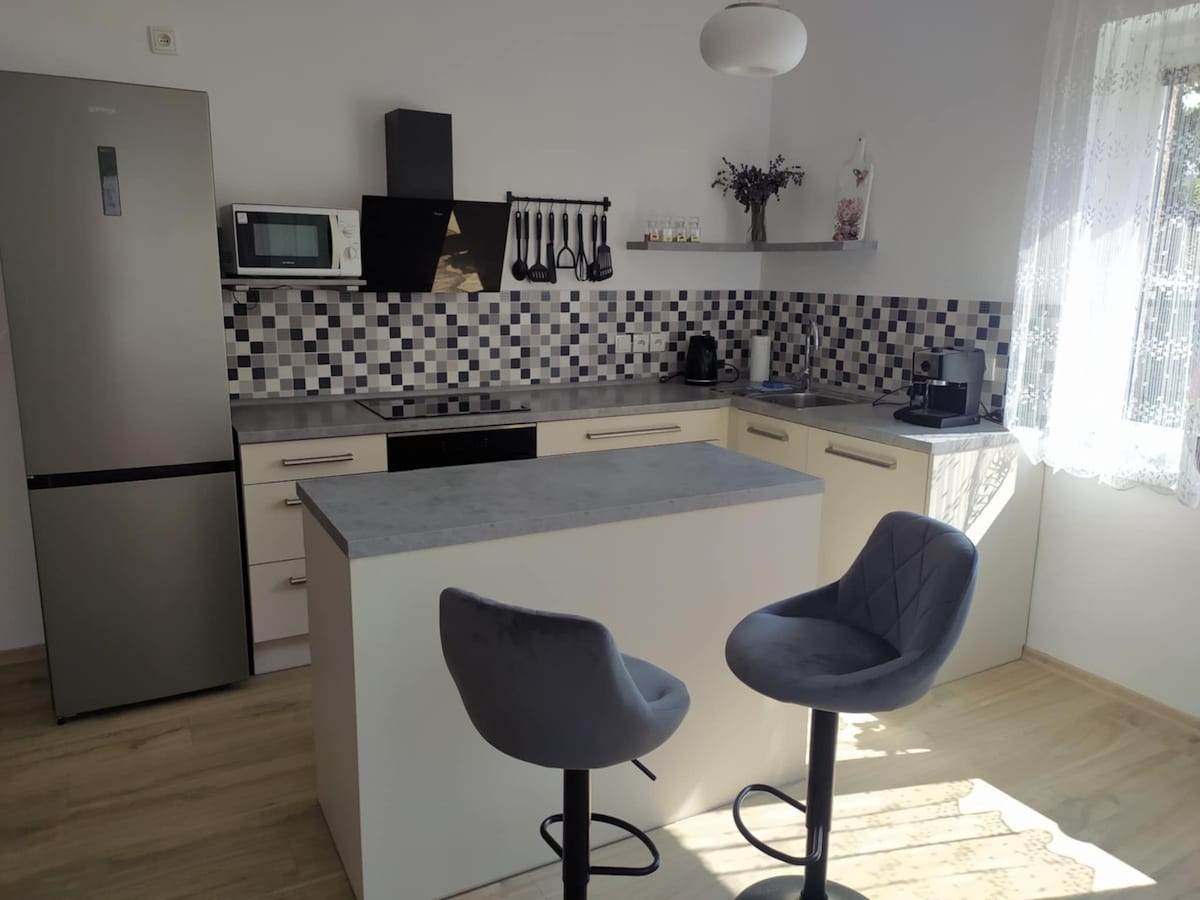 A modern kitchen is equipped with a sleek stainless-steel refrigerator and a microwave. Black and white tiles create a visually appealing backsplash. Bar stools are positioned near the counter, which features a smooth surface. Natural light filters in through a window adorned with lace curtains.
