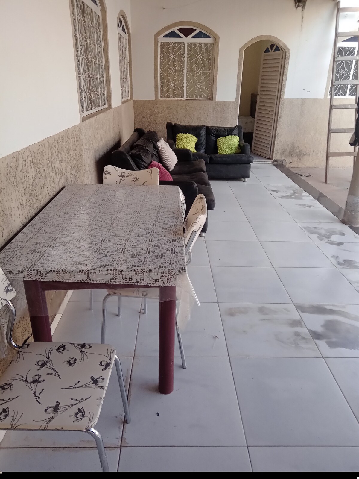 An outdoor area showcases a long table with patterned tablecloth, accompanied by several chairs. In the background, a collection of dark sofas adorned with colorful cushions provides a cozy seating arrangement. Natural light brightens the space, highlighting the tiled floor.