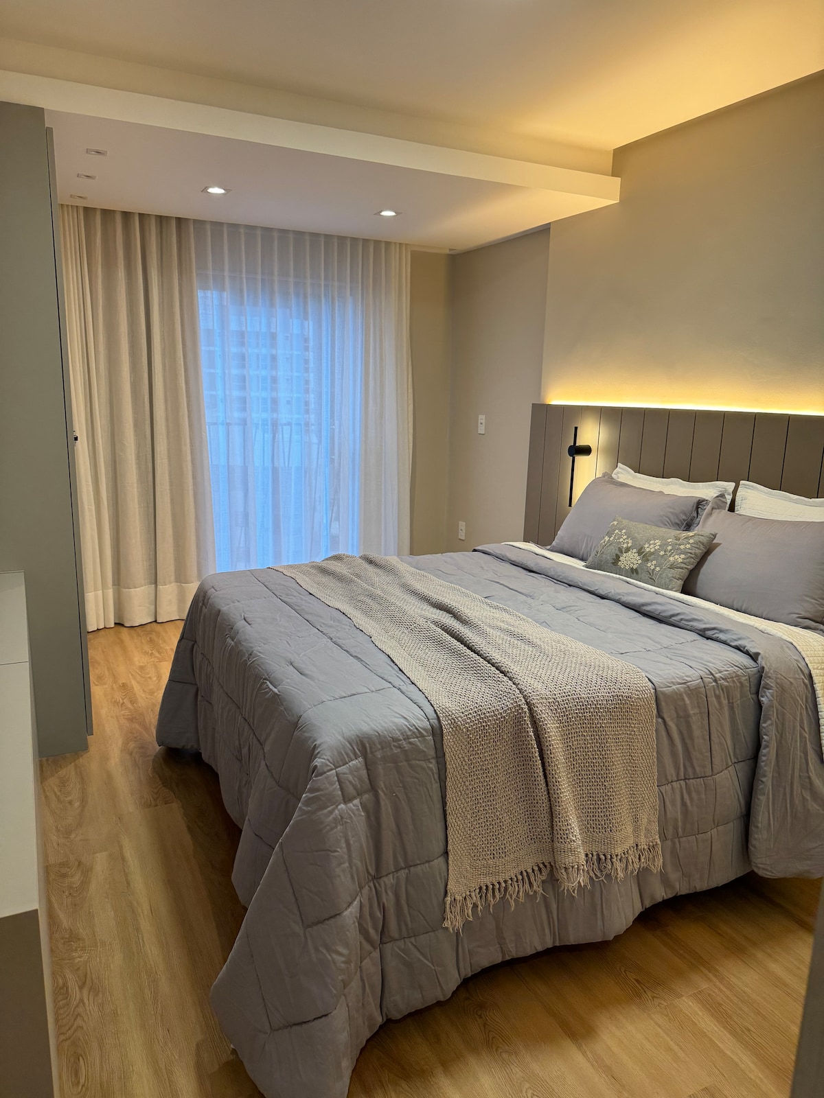 A calming bedroom features a spacious bed covered with a soft grey comforter. Natural light filters through sheer curtains next to a glass door leading to a balcony. Subtle lighting highlights the textured wall behind the headboard, creating a serene atmosphere.