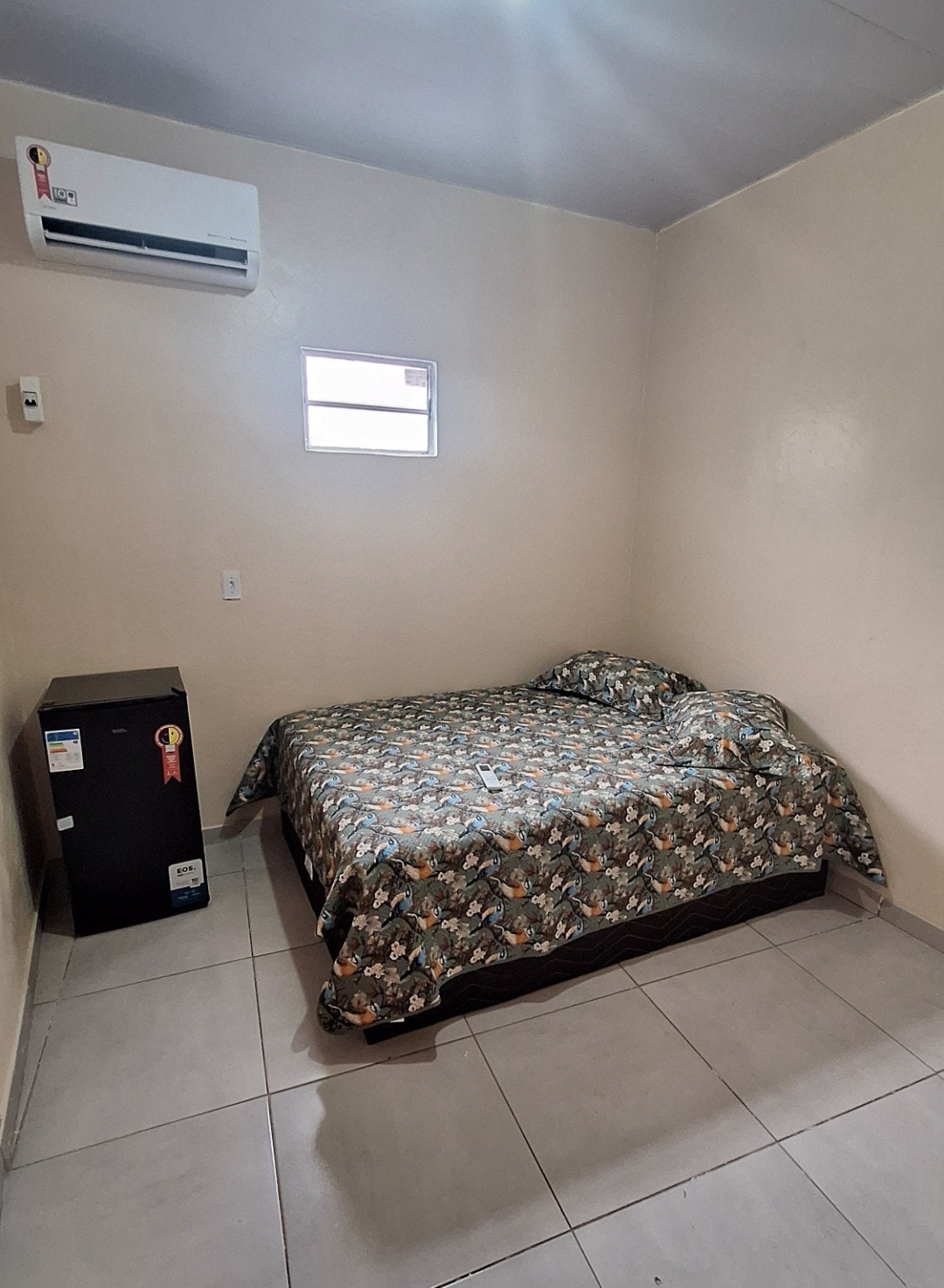 A simple sleeping area is furnished with a spacious bed covered in a patterned blanket. A small refrigerator is positioned nearby, and a window allows natural light to enter the room. The walls are painted in a neutral tone, creating a calm atmosphere.