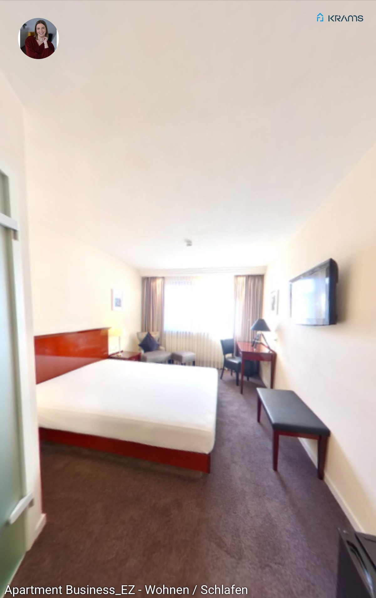 A spacious room is presented with a large, unmade bed and a mounted television. A desk and chair are positioned across from the bed. Natural light enters through a large window, partially covered by curtains, while neutral wall tones enhance the room's calm ambiance.