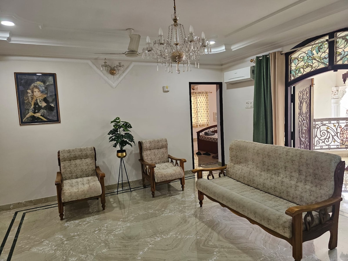 The living area is furnished with a three-seat sofa and two armchairs, all featuring wooden armrests. A plant adds a touch of greenery to the room. Natural light enters through a door leading to a bedroom, while a decorative chandelier hangs from the ceiling.