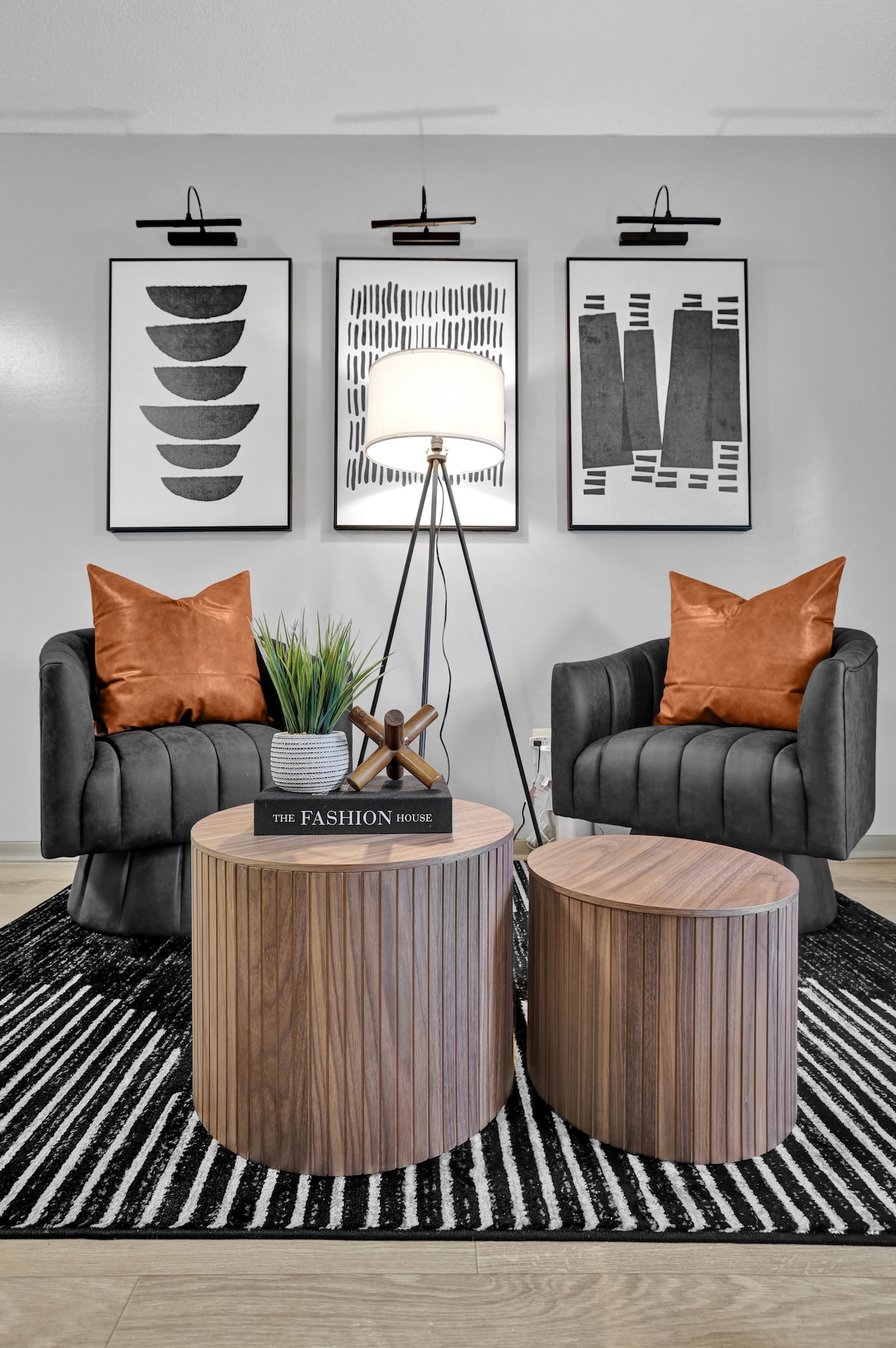 A modern seating area features two curved black chairs with orange accent pillows, arranged around a round wooden coffee table. A secondary, smaller side table is nearby. The area is enhanced by patterned wall art and a tall lamp, all set against a striped rug.