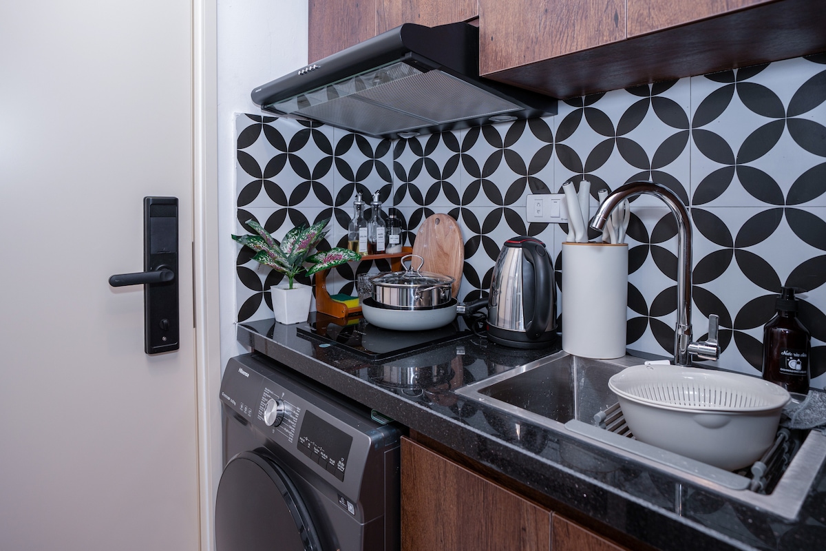 A modern kitchen is equipped with a stainless steel sink, a compact stove, and a variety of kitchenware, including pots and utensils. A washing machine is positioned below the counter. The wall features a geometric black-and-white tile design, complemented by a small potted plant.