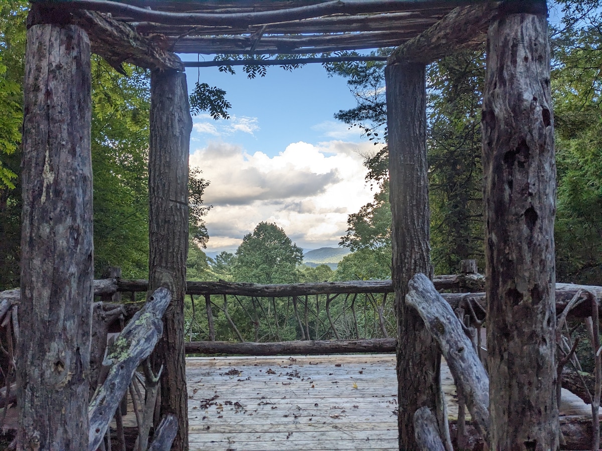 A wooden frame constructed from natural logs creates an open viewpoint overlooking rolling hills and distant mountains. Lush greenery surrounds the structure, while fluffy clouds drift in a clear blue sky, emphasizing the tranquility of the setting.