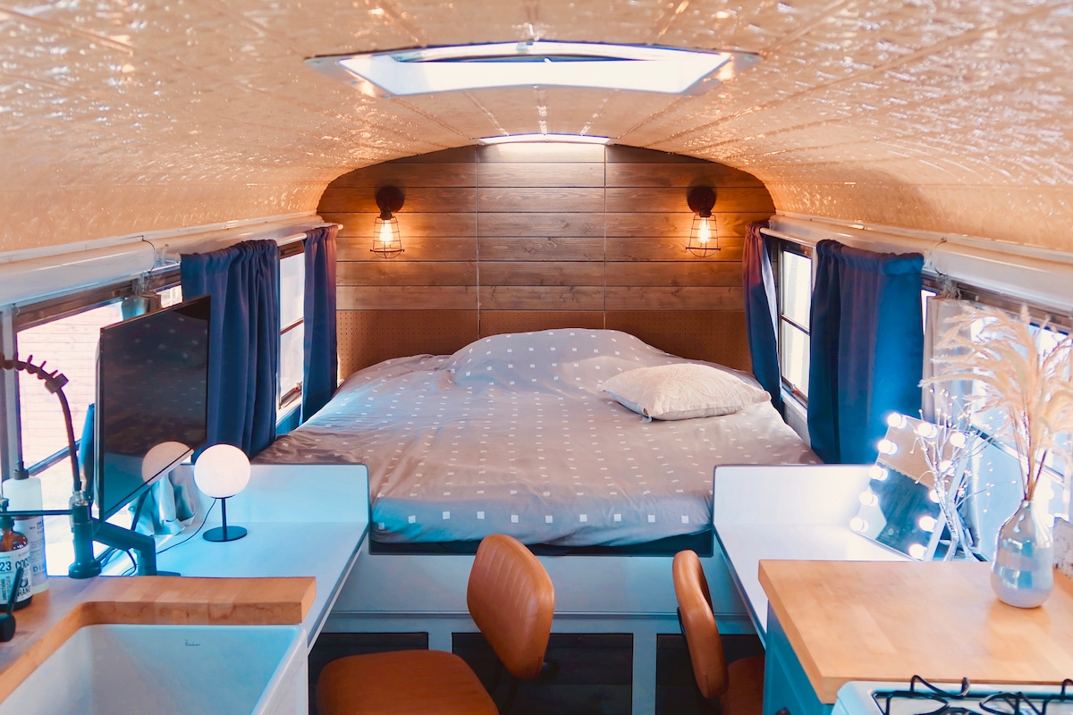 An interior view of a cozy camper is presented, featuring a spacious bed with a dotted gray comforter. Warm wood paneling adorns the walls, while large windows draped with dark curtains allow natural light to enter. A small dining area with two leather chairs and a wooden table is visible.