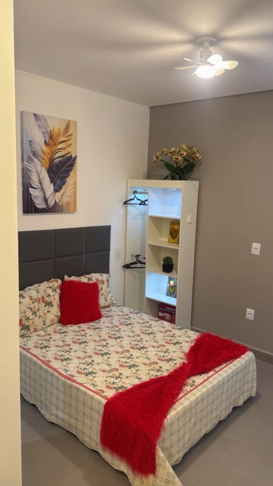 A cozy bedroom features a spacious bed adorned with a floral blanket and red accents. A decorative wall piece of tropical leaves is displayed above the bed. A shelving unit is positioned nearby, displaying ornamental items and providing additional storage.