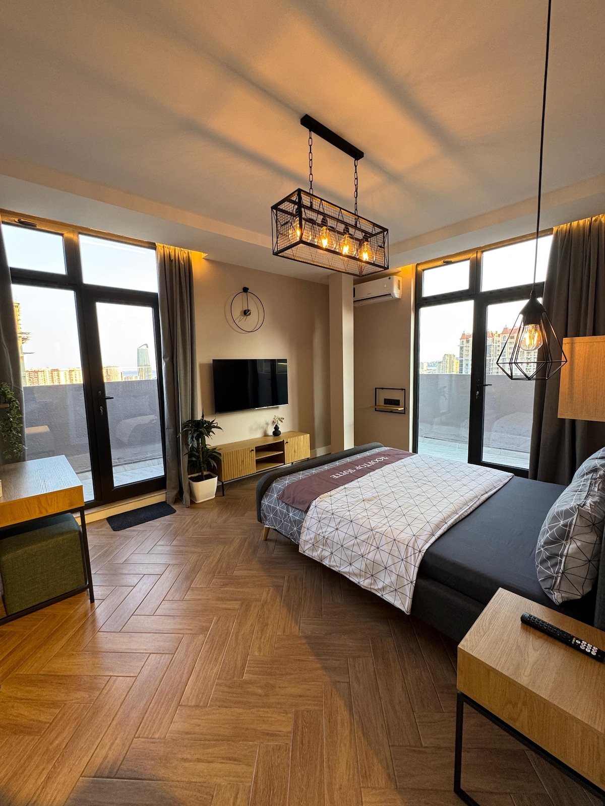 A well-lit bedroom features large windows that offer city views and allow natural light to fill the space. A comfortable bed with an elegant duvet is paired with a contemporary bedside table. A modern light fixture hangs above, and a TV is mounted on the wall.