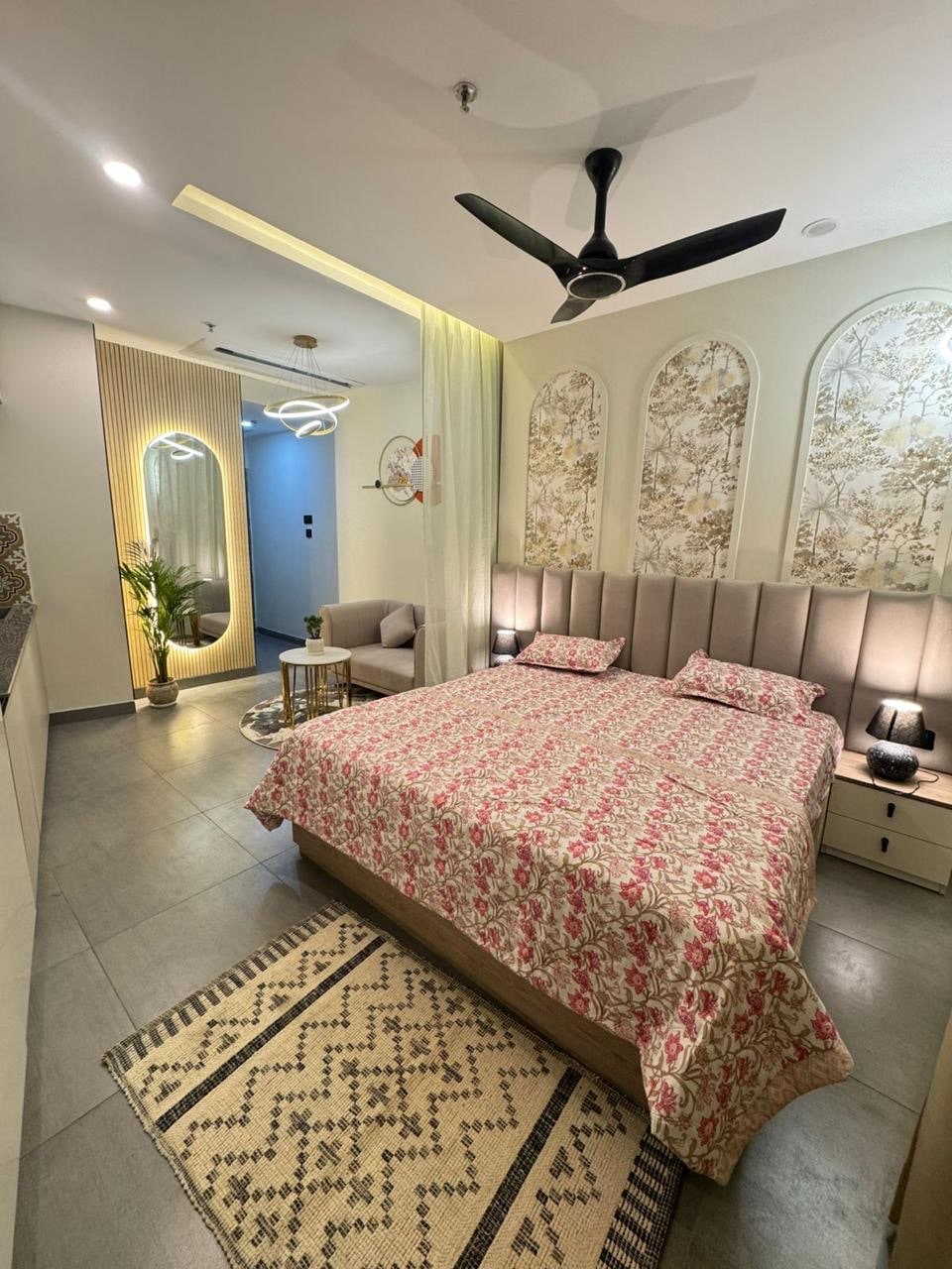 A spacious bedroom featuring a comfortable bed dressed in floral bedding. Decorative arches adorn the wall, and soft lighting enhances the ambiance. A cozy seating area with a small table is visible, along with a patterned area rug on the floor.