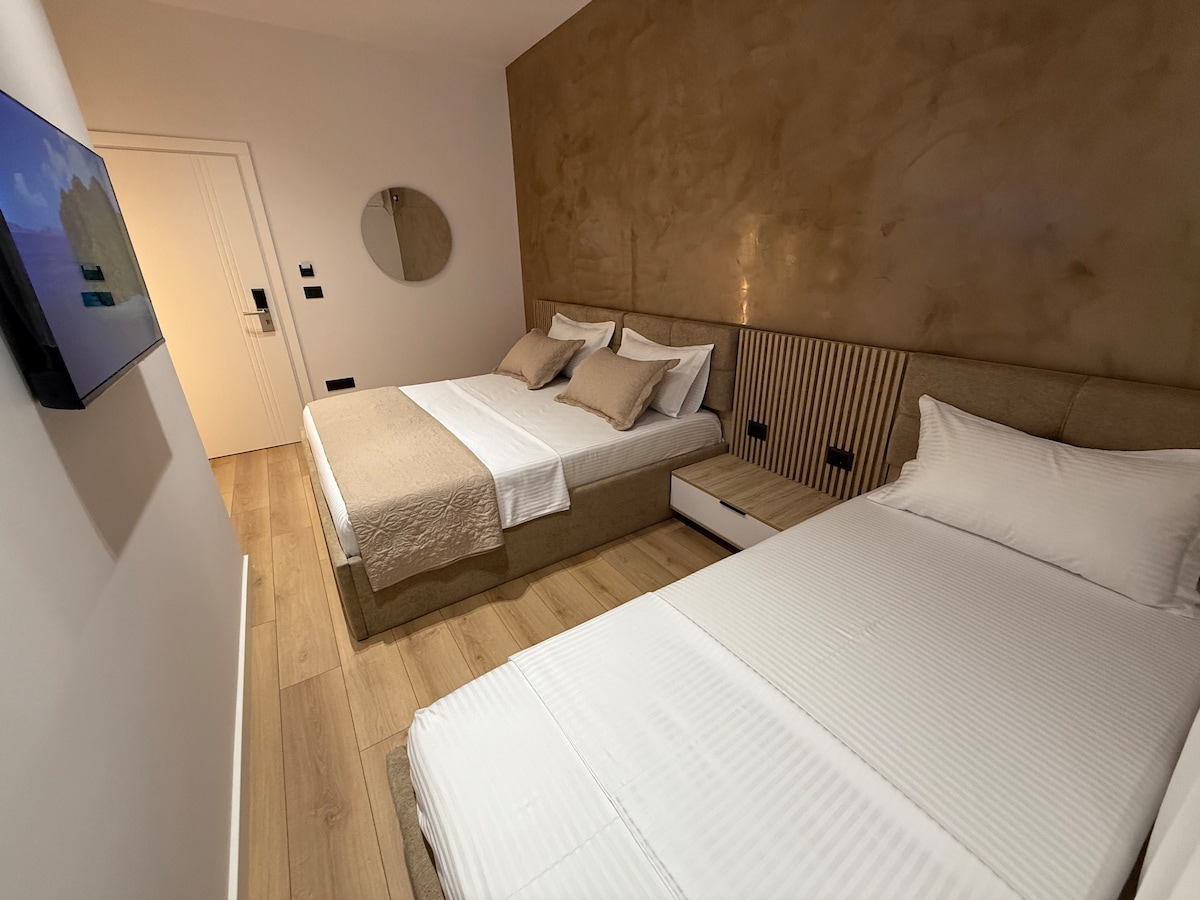 A modern guest room features two spacious beds with neutral-toned linens. A subtle textured wall adds warmth to the space. A round mirror and flat-screen TV are mounted on the wall, while light wooden flooring completes the contemporary decor.