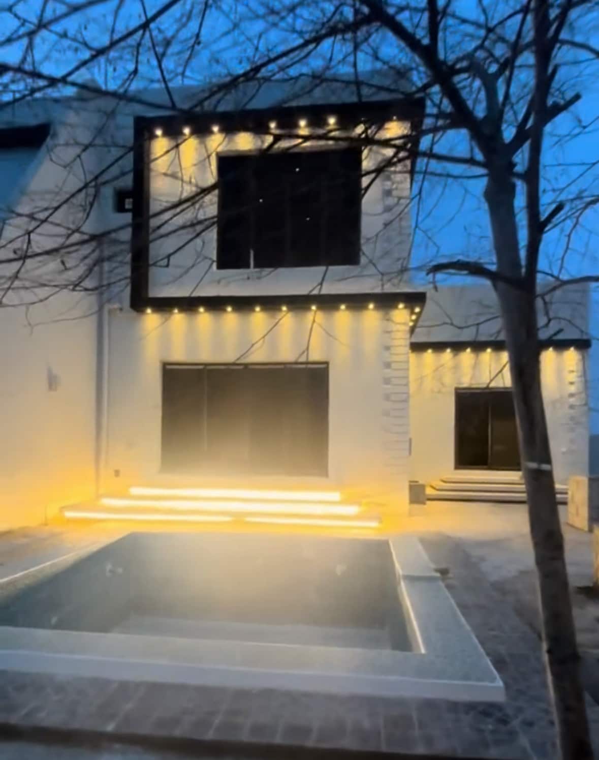The exterior of a modern home is illuminated by soft lighting at dusk. A small pool is situated in front, complemented by steps leading up to the entrance. Large windows reflect the warm glow, surrounded by minimal landscaping.