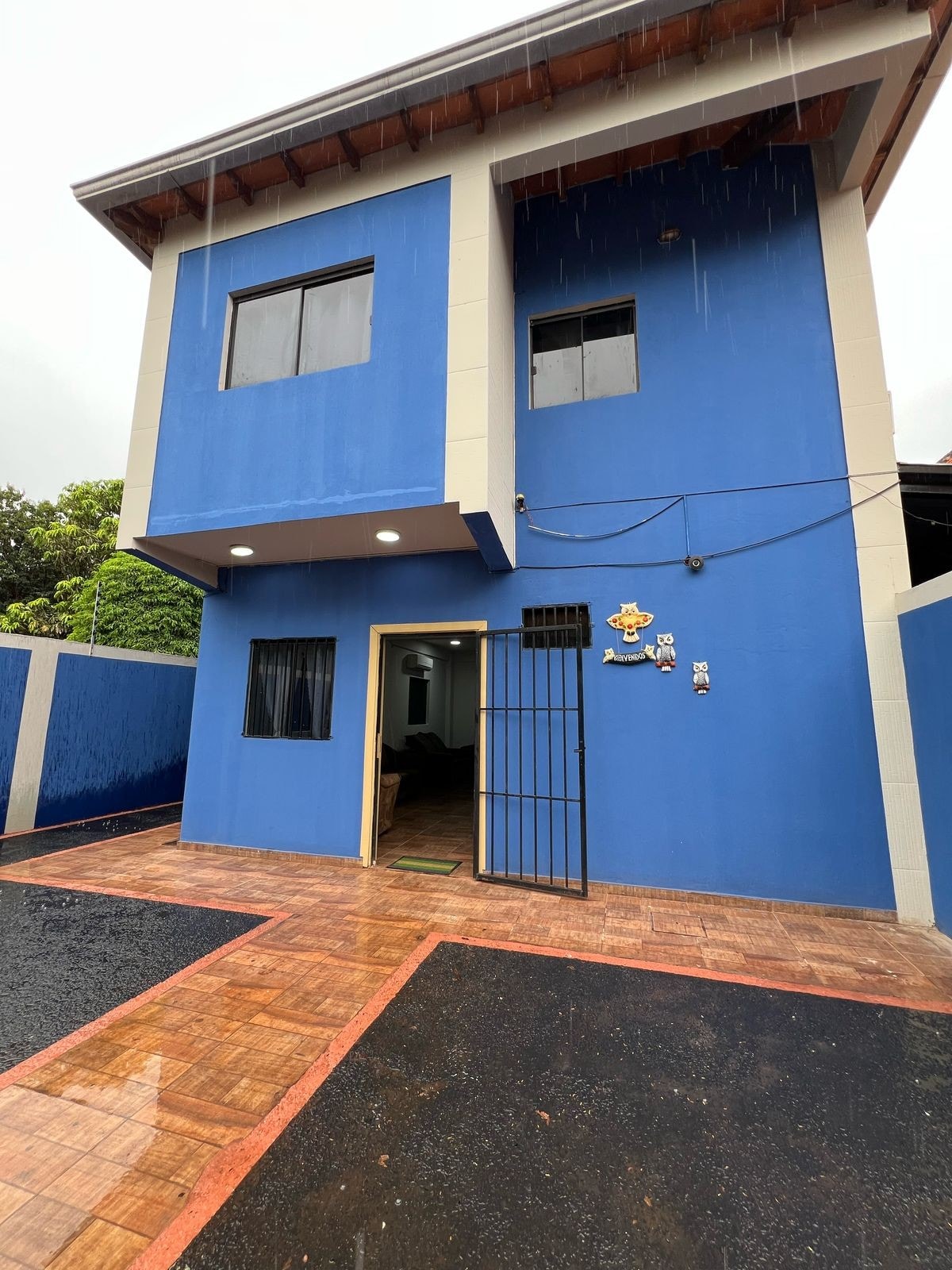 An exterior view of a two-story blue house is presented, featuring multiple windows and a metal gate. A paved area is visible in front, with a polished surface showing reflections from rain. Lush greenery is seen in the background, alongside a welcoming entrance.