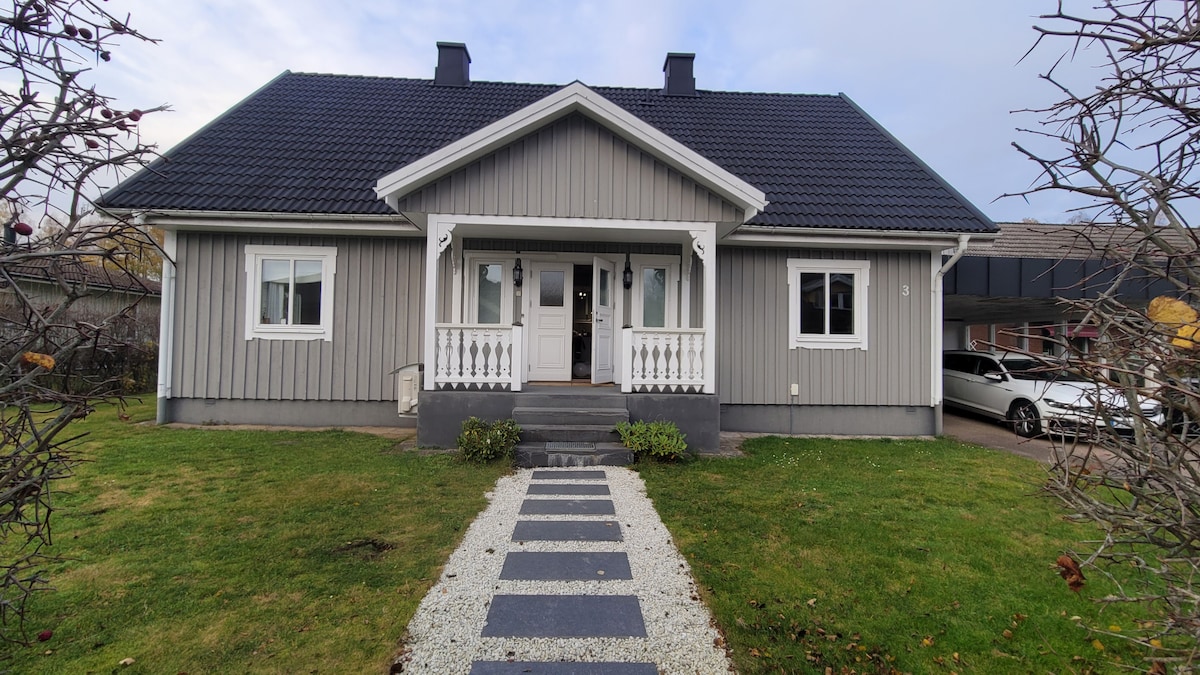 The exterior of a spacious family home is presented, featuring a light gray facade and a welcoming porch with decorative railing. A stone pathway leads through a well-maintained garden, flanked by greenery. A driveway with parked vehicles is visible to the side.