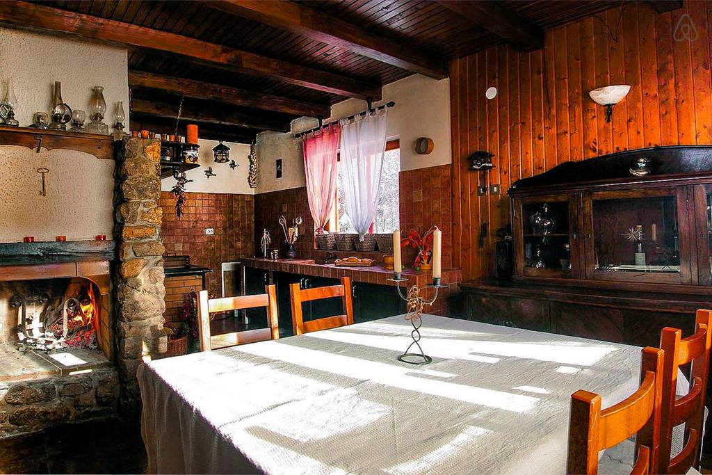 A rustic dining area features a large wooden table surrounded by several chairs. Natural light filters through the partially draped windows, illuminating the stone fireplace and wooden cabinetry. A cozy atmosphere is enhanced by wooden beams overhead and warm-toned walls.