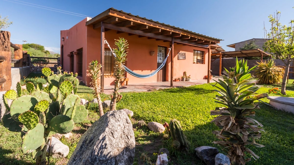 A single-story house is showcased amidst lush green landscaping, featuring various plants and rocks. A hammock hangs on the porch beneath a wooden overhang, offering a spot for relaxation. The exterior walls are painted in a warm peach color, complementing the natural surroundings.
