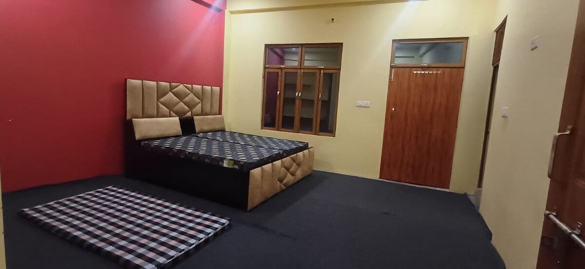 A spacious bedroom is presented with a large bed featuring a geometric-patterned headboard. A complementary mat is placed on the floor. The room is illuminated by natural light from multiple windows, while walls painted in red and yellow create a vibrant atmosphere.