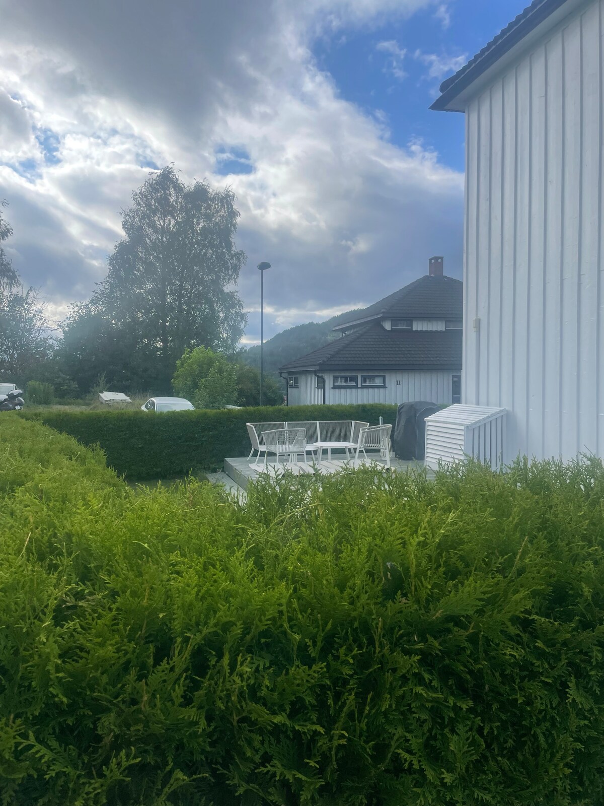 An outdoor dining area is surrounded by lush greenery and features a white table with matching chairs. The scene is complemented by a cloudy sky above and a glimpse of the house in the background.