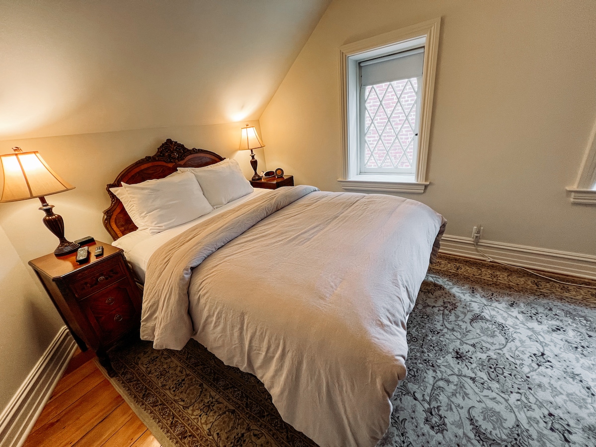 A queen-sized bed is centered in a softly lit room, flanked by two bedside lamps on elegant wooden tables. A window with diamond-patterned glass is positioned to the right, providing natural light. A decorative area rug covers the hardwood floor beneath.