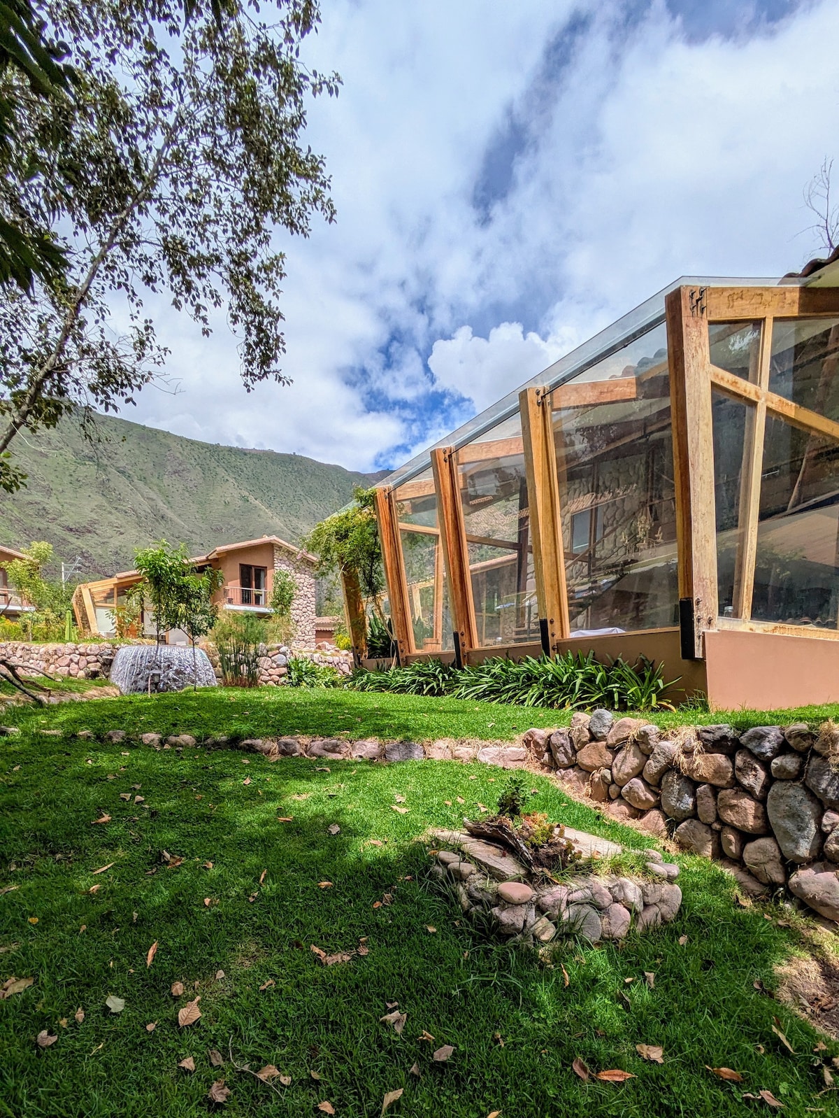 A serene outdoor view features a glass-walled structure surrounded by lush greenery. The landscape is complemented by stone pathways and flower beds, with mountains rising in the background under a partly cloudy sky.