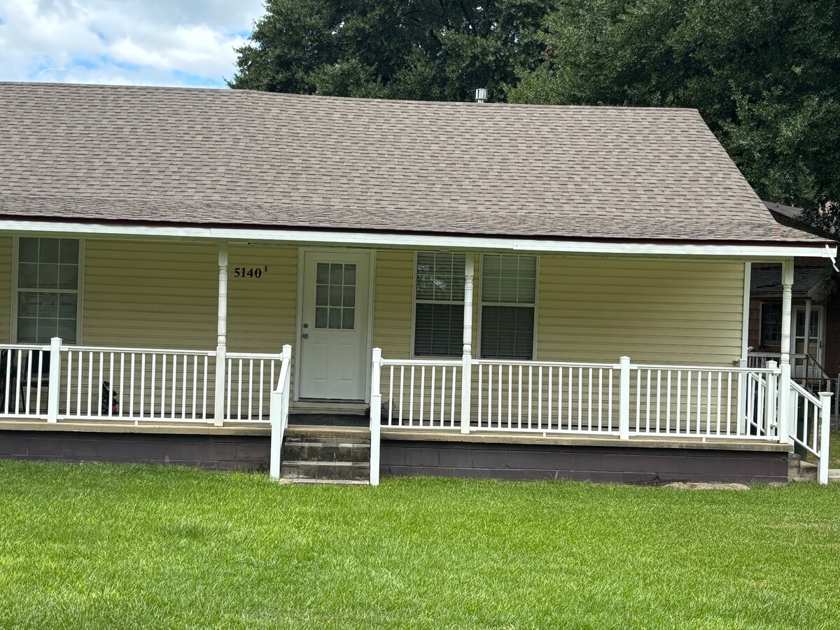 A single-story home with a welcoming front porch features white railing. Large windows provide natural light to the interior. A grassy lawn surrounds the house, and a concrete pathway leads to the front door, creating an accessible entrance.