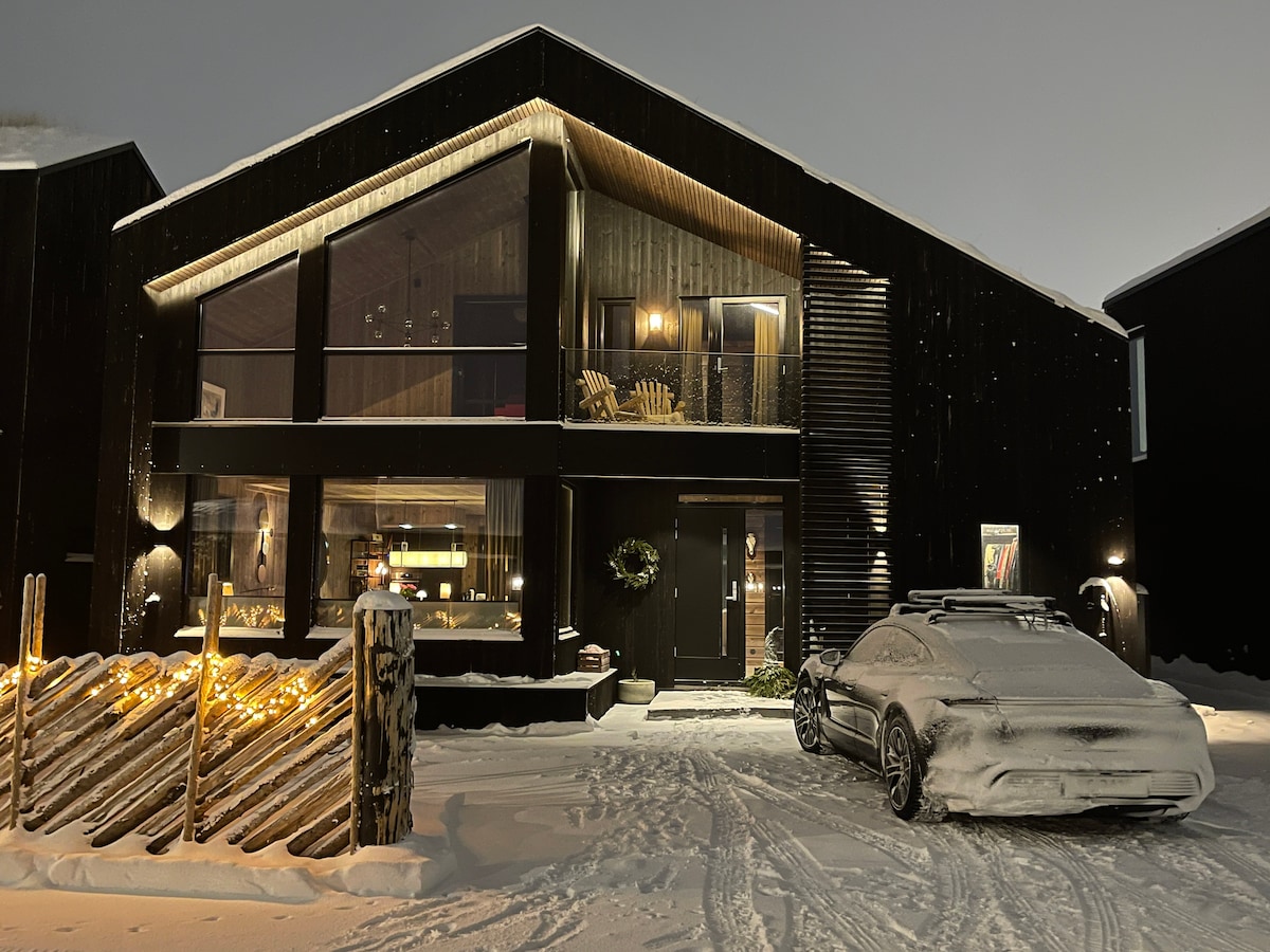 The exterior of a modern black chalet is presented, featuring large glass windows that reflect the surrounding snowy landscape. A car is parked in front, and a wooden fence is decorated with warm lights. Soft snow blankets the ground, adding to the serene winter ambiance.