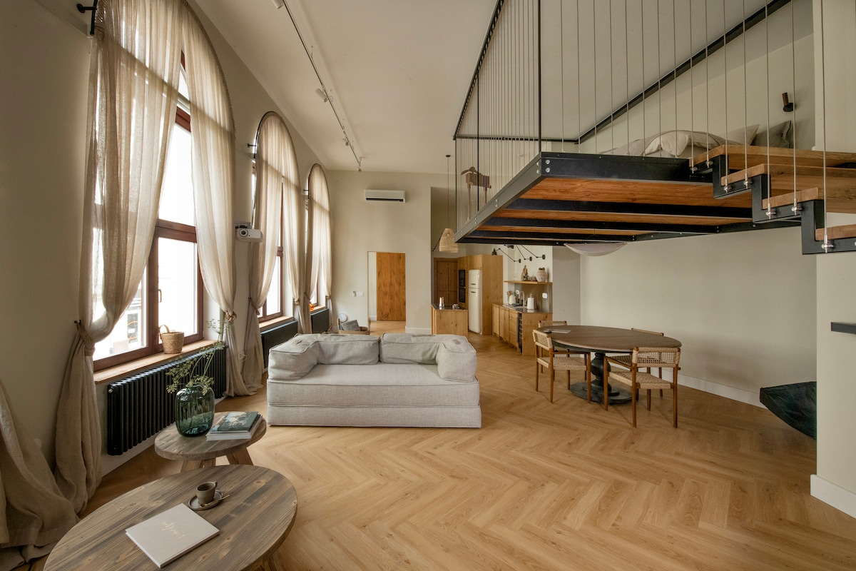 An open living area features large windows dressed with sheer curtains, allowing natural light to fill the space. A spacious sofa is positioned near a round wooden dining table, surrounded by chairs. A lofted sleeping area can be seen elevated above the kitchen area.