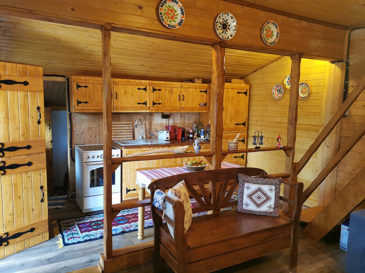 Inside a rustic cabin, wooden features dominate, including a seating area with a simple couch. A well-equipped kitchen is visible, with wooden cabinets and a stove. Decorative plates hang on the walls, and traditional textile accents enhance the cozy environment.